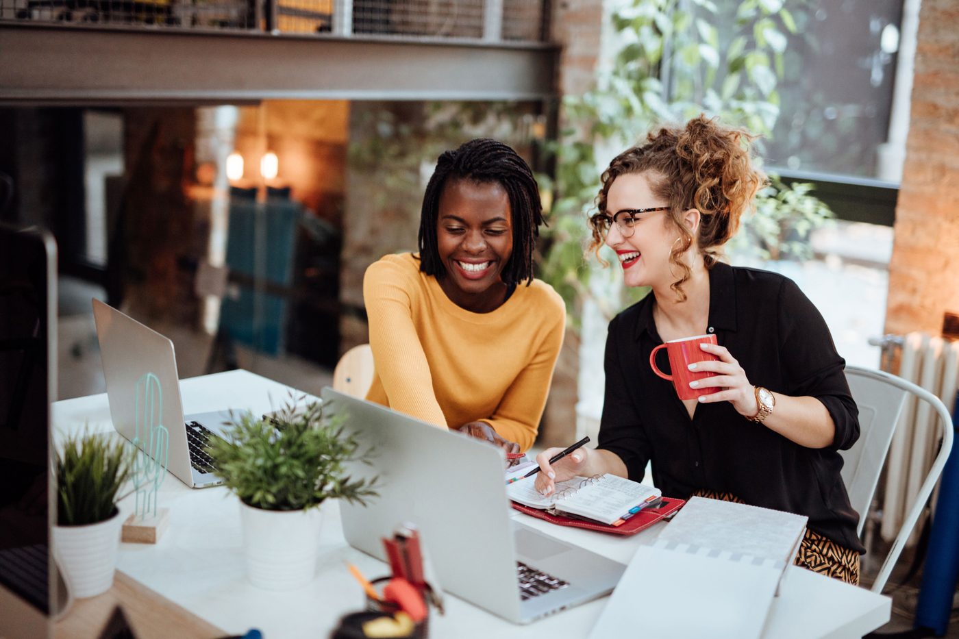 Two women laughing over a laptop at a shared table